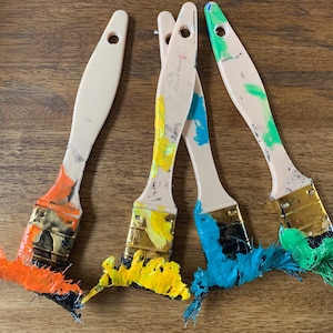 May include: Four paint brushes with wooden handles and gold metal ferrules. The brushes are covered in dried paint in various colors, including orange, yellow, blue, and green.