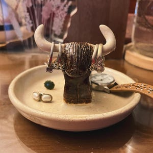 Puede incluir: Un porta anillos de cerámica con forma de vaca de las Highlands con pelaje marrón y negro. El soporte tiene un plato de cerámica blanco con un reloj y anillos encima.