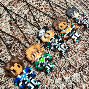 May include: Five pixel art pendants made from small plastic beads, each featuring a different character design with distinct hair and clothing colours, including blue, green, black, orange, and red accents, all attached to thin black cords.