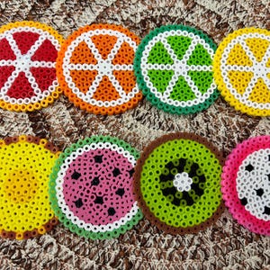 Pode incluir: Oito porta-copos circulares feitos de contas de plástico coloridas, dispostos em duas filas. Os designs apresentam vários padrões de frutas, incluindo fatias de citrinos vermelhas, laranjas, verdes e amarelas, um ananás, melancia, kiwi e pitaia.