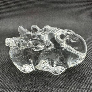 May include: A clear, solid glass sculpture featuring an abstract, organic shape with multiple rounded protrusions and smooth, flowing contours. The piece is transparent, reflecting light across its uneven, polished surface, creating a complex form.