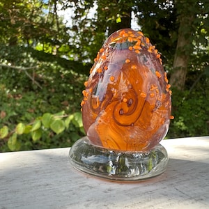 May include: An egg-shaped glass sculpture in shades of orange and brown, with decorative orange dots. The sculpture sits on a clear, oval glass base. The background features green foliage. The sculpture's dimensions are approximately 15 cm in height.