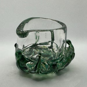 May include: A clear glass bowl with a unique design. The bowl has a cylindrical shape with a rounded top and is supported by a base of swirling green glass. The green glass base has a decorative, organic shape.