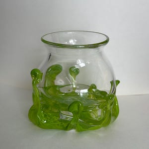 May include: A clear glass bowl featuring a wide, rounded body and a flared rim. The base is adorned with organic, swirling lime green glass accents that extend upward from the bottom, creating a textured, fluid appearance around the exterior.