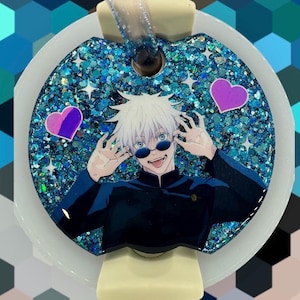 May include: A decorative car air freshener featuring a cartoon character with white hair and sunglasses. The air freshener has a glittery blue background with star accents and two pink hearts. The air freshener is attached to a white and beige plastic base.