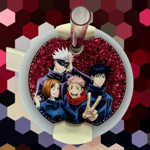May include: A red glittery drink charm featuring a group of anime characters. The charm has a white border and a clear straw. The background is a geometric pattern of red and purple hexagons.