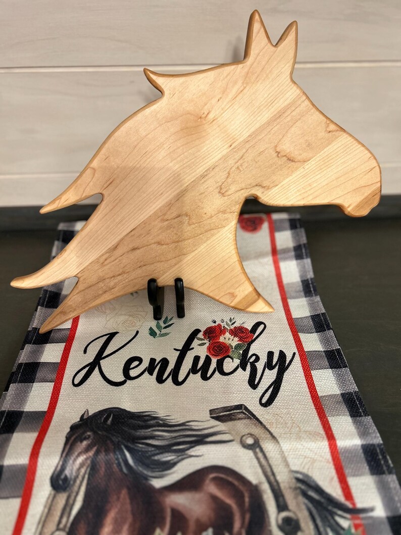 May include: A wooden horse head-shaped chopping board is displayed on a black stand. The board is light brown with visible wood grain. Below, a decorative runner features the word "Kentucky" and a horse illustration.