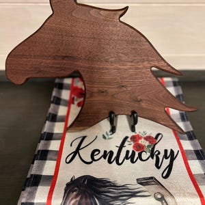 May include: A dark brown wooden horse head-shaped towel holder with two black hooks. The holder is over a towel with a black and white plaid pattern, red accents, and the word "Kentucky" in script.