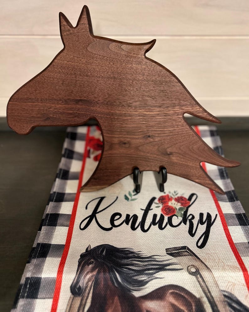 May include: A dark brown wooden horse head-shaped towel holder with two black hooks. The holder is over a towel with a black and white plaid pattern, red accents, and the word "Kentucky" in script.