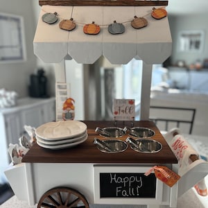 May include: A white and brown decorative cart with a wooden wheel, a chalkboard sign that says "Happy Fall!", and a garland of pumpkin cutouts. The cart is decorated with fall-themed items, including pumpkins and signs.