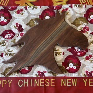 May include: A wooden horse-head shaped chopping board. The board is a dark brown with lighter wood grain. The background has a festive pattern of red and white ornaments, with the text "HAPPY CHINESE NEW YEAR".