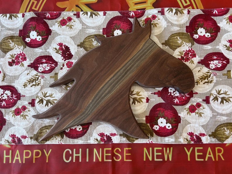 May include: A wooden horse-head shaped chopping board. The board is a dark brown with lighter wood grain. The background has a festive pattern of red and white ornaments, with the text "HAPPY CHINESE NEW YEAR".