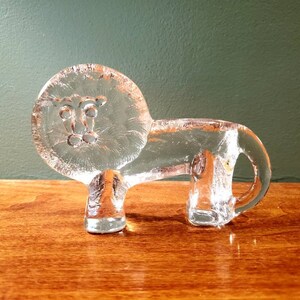 May include: A clear glass lion figurine with a detailed face. The lion is standing on a wooden surface.