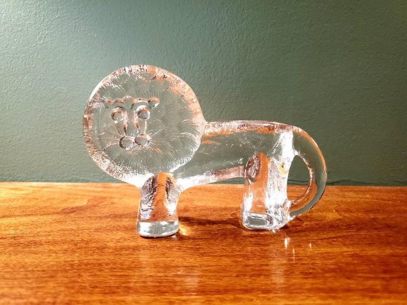 May include: A clear glass lion figurine with a detailed face. The lion is standing on a wooden surface.