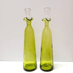 May include: Two green glass bottles with a ruffled top and a clear glass stopper. The bottles are on a white surface.