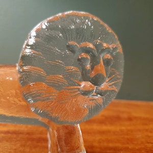 May include: A glass lion figurine with a detailed face and mane. The lion is standing on a wooden surface.