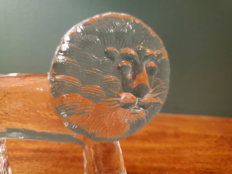 May include: A glass lion figurine with a detailed face and mane. The lion is standing on a wooden surface.