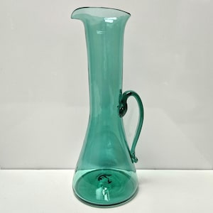 May include: A tall, teal-colored glass pitcher with a flared top and a curved handle. The pitcher has a smooth surface and a rounded base. The handle is attached near the top and bottom of the pitcher. The pitcher is empty and sits on a white surface.