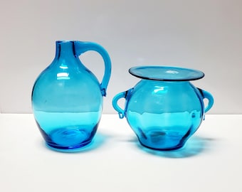 May include: Two turquoise glass vessels. One is a pitcher with a handle, the other is a round bowl with two small handles and a flared rim. Both are transparent and sit on a white surface.