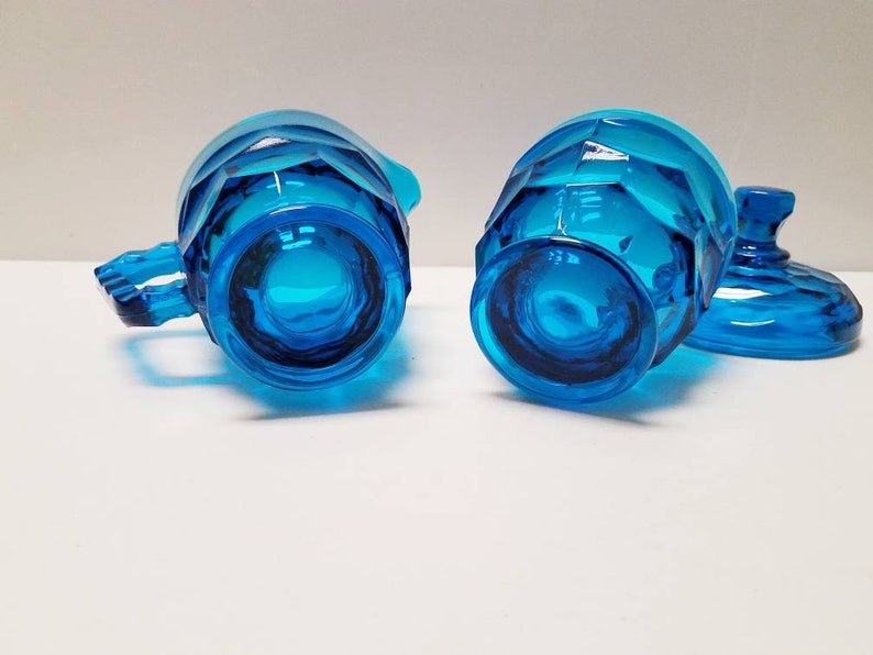 May include: A vintage blue glass creamer and sugar bowl set with a faceted design. The creamer has a handle and the sugar bowl has a lid.