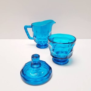 May include: A blue glass sugar bowl with a lid and a matching creamer. The sugar bowl and creamer have a faceted design.