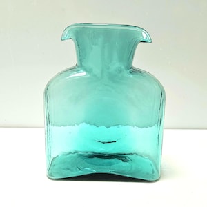 May include: A translucent teal glass vase with a rectangular body and a wide, flared neck. The glass features a smooth, slightly wavy texture throughout its surface, with a distinct curved rim that extends outward on both sides of the opening.