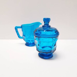May include: A blue glass creamer and sugar bowl set with a geometric pattern. The creamer has a handle and the sugar bowl has a lid.
