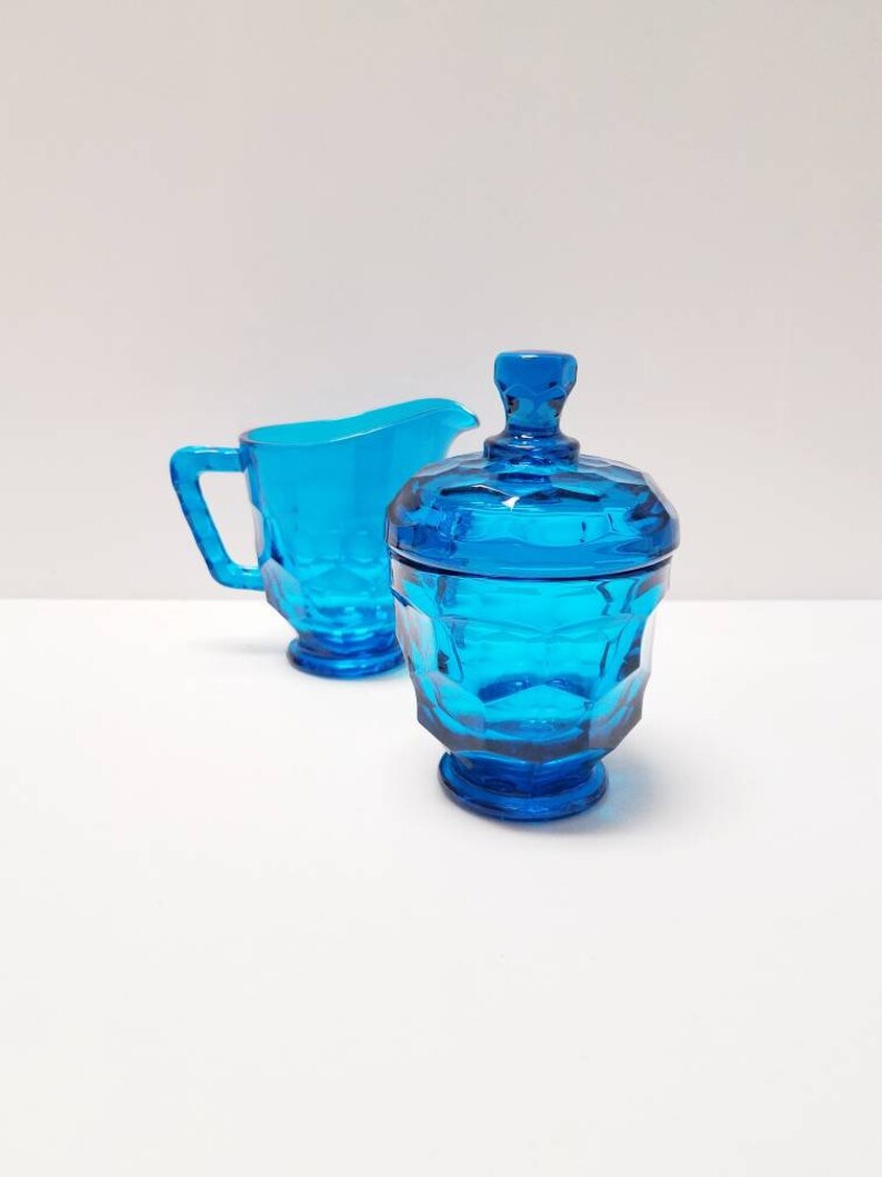 May include: A blue glass creamer and sugar bowl set with a geometric pattern. The creamer has a handle and the sugar bowl has a lid.