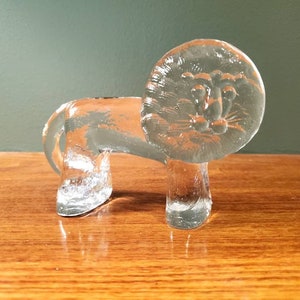 May include: A clear glass lion figurine with a detailed face and a round head. The figurine is standing on a wooden surface.
