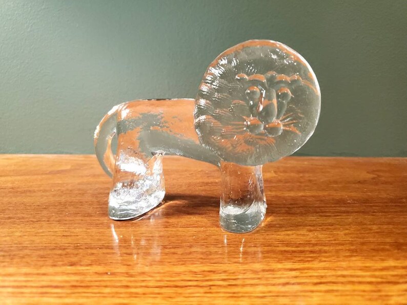 May include: A clear glass lion figurine with a detailed face and a round head. The figurine is standing on a wooden surface.