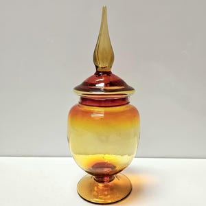 May include: A decorative glass jar with a lid and a pointed finial. The jar transitions from a golden yellow to a reddish-orange hue. The jar sits on a round base.