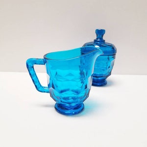 May include: A blue glass creamer and sugar bowl set. The creamer has a handle and a rounded base. The sugar bowl has a lid and a round base.