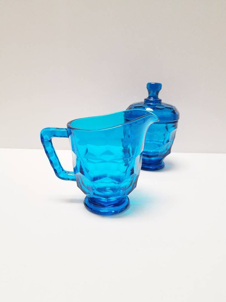 May include: A blue glass creamer and sugar bowl set. The creamer has a handle and a rounded base. The sugar bowl has a lid and a round base.