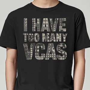May include: Black t-shirt with the text "I HAVE TOO MANY VCAS" in a dotted pattern. The text is in a light grey color, contrasting with the dark background. The shirt is a casual wear item.