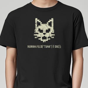 May include: Black t-shirt featuring a pixelated cat face design in off-white. Below the cat graphic, the text reads "HUMAN.FEED("TUNA") || DIE();". The shirt is made of a soft material.