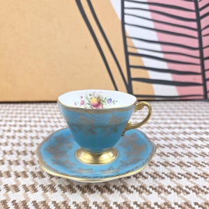 Puede incluir: Un juego de taza y platillo de té turquesa vintage con detalles dorados. La taza presenta un diseño floral en el interior del borde y un asa dorada. El platillo y la taza tienen patrones de filigrana dorados. El juego está sobre una superficie estampada.