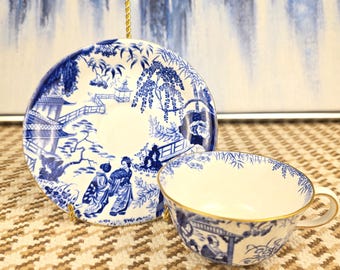 May include: A blue and white porcelain teacup and saucer set. The set features a detailed Asian-inspired landscape design. The teacup has a gold rim. The saucer is propped up by a gold stand.