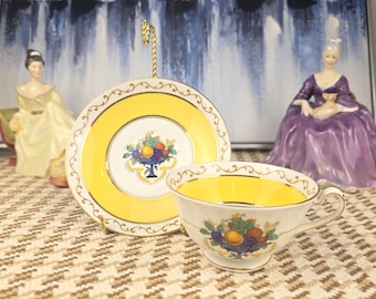 May include: A vintage teacup and saucer set with a yellow interior and exterior, adorned with a fruit basket design. The set includes a matching saucer and a teacup with a gold trim. Decorative figurines are in the background.
