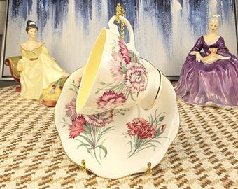 May include: A vintage teacup and saucer set with a floral design featuring pink carnations and green leaves. The cup has a yellow interior and gold trim. The saucer is scalloped with a matching floral pattern. The set is displayed on a patterned surface.