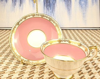 May include: A pink teacup and saucer set with gold trim and floral detailing. The teacup has a white exterior and a pink interior. The saucer is pink with a white center. The set is on a patterned surface.