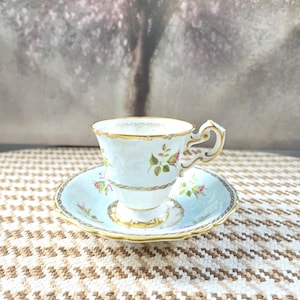 May include: A porcelain teacup and saucer set featuring a pale blue background with delicate pink rosebud motifs, green leaves, and ornate gold trim along the scalloped edges and the handle. The cup has a pedestal base and a decorative handle.