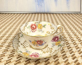 Puede incluir: Un juego de taza y platillo de té vintage. La taza y el platillo son de color crema con ribetes dorados y diseños florales en rojo, amarillo y azul. El juego está colocado sobre una superficie estampada.