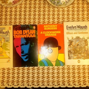 Peut inclure: Quatre livres de poche vintage sont présentés sur une surface à motifs. Les titres incluent "The Loved One" d'Evelyn Waugh, "Tarantula" de Bob Dylan, "A Clockwork Orange" d'Anthony Burgess et "Officers and Gentlemen" d'Evelyn Waugh.