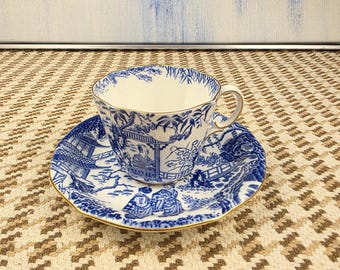 May include: A blue and white porcelain teacup and saucer set. The cup and saucer feature a detailed landscape design with gold trim. The set is placed on a patterned beige and brown surface, with a blue and white abstract painting in the background.