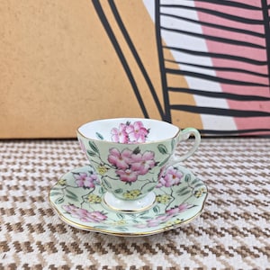 May include: A vintage teacup and saucer set. The cup and saucer are light green with pink floral patterns and gold trim. The teacup has a small handle. The set is placed on a patterned surface.