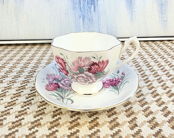 May include: A white teacup and saucer set with a floral design in shades of pink, purple, and green. The teacup and saucer have gold trim. The set is placed on a patterned surface.