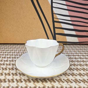 May include: A white teacup and saucer set with gold trim. The teacup has a scalloped rim and a gold handle. The saucer has a floral design. The set is on a patterned surface.
