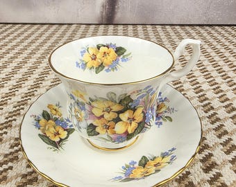 May include: A white teacup and saucer set with a floral design featuring yellow and blue flowers. The cup and saucer have gold trim. The teacup has a curved handle.