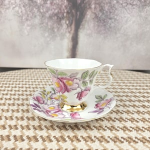 May include: A fine bone china teacup and saucer set. The white cup and saucer are decorated with pink and purple floral patterns and green leaves. The cup has a gold handle and base. The saucer is round. The set is on a patterned surface.