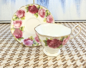 May include: A vintage teacup and saucer set. The white porcelain cup and saucer are decorated with pink and burgundy roses and gold trim. The teacup has a pedestal base and a curved handle. The saucer is round with a scalloped edge.